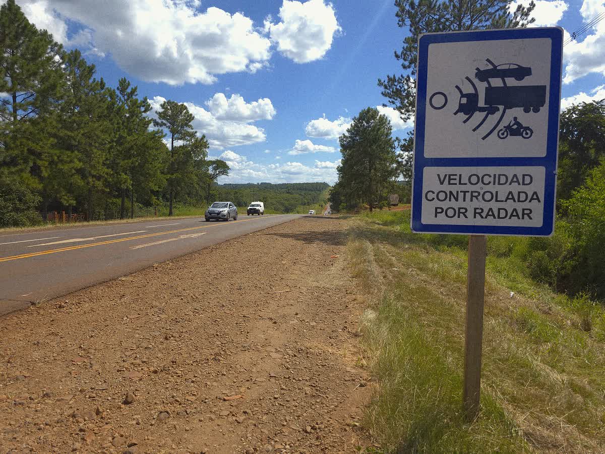 Cartel vertical al borde de una carretera con el pictograma de un radar apuntando a un auto, un camión y una moto, y el texto VELOCIDAD CONTROLADA POR RADAR